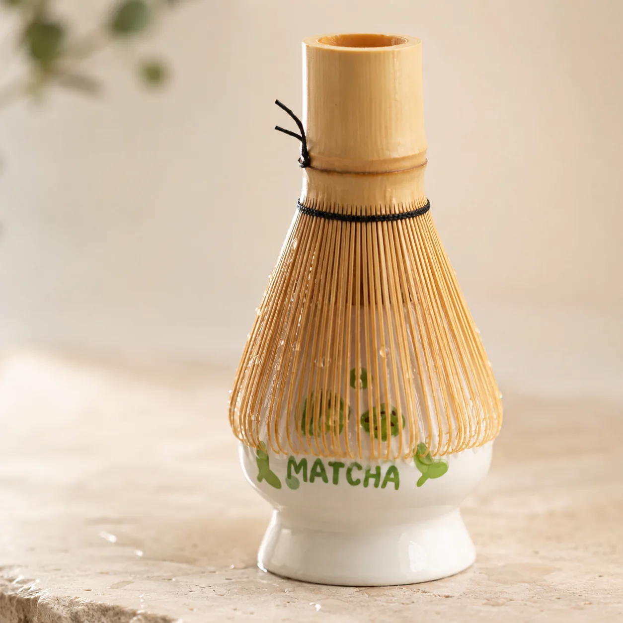 Close-up of a freshly rinsed bamboo chasen resting on a white glazed ceramic holder painted with green matcha lettering, water droplets clinging to the tines